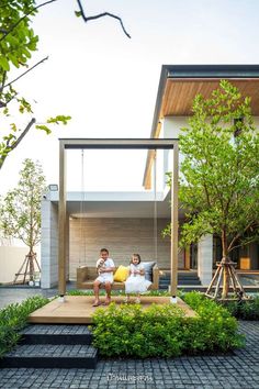 two children sitting on a swing in front of a house with trees and bushes around them