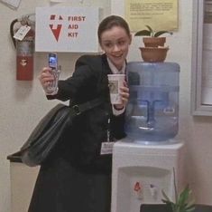 a woman standing next to a water dispenser holding a cup and cell phone