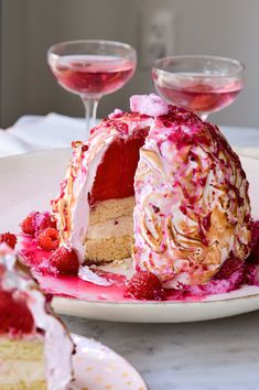 a cake with white frosting and raspberry toppings on a plate next to two wine glasses