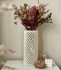 a crocheted vase with dried flowers in it on a white table next to two balls of yarn