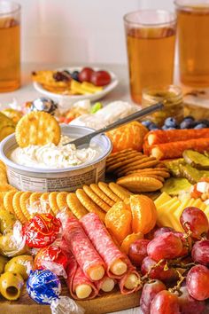 an assortment of cheeses, crackers and fruit on a platter with glasses of beer in the background