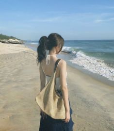 a woman standing on top of a sandy beach next to the ocean holding a purse