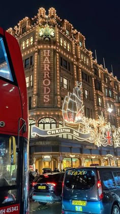 a red double decker bus driving past a tall building with christmas lights on it's side