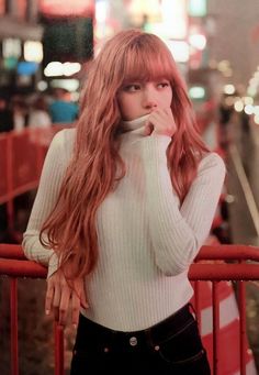 a woman with long red hair standing next to a fence in front of a city street