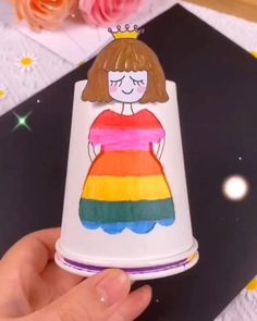 a hand holding a paper cup with a drawing of a girl wearing a rainbow dress