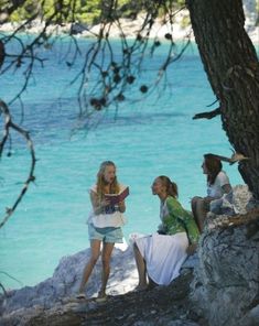 three women sitting on rocks near the water and talking to each other while one looks at her cell phone