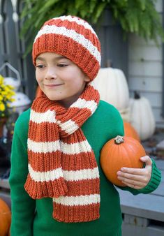 a young boy wearing a knitted hat and scarf holding two pumpkins in his hands