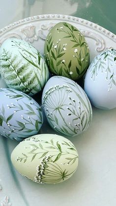 four painted eggs sitting on top of a white plate with green leaves and flowers in them