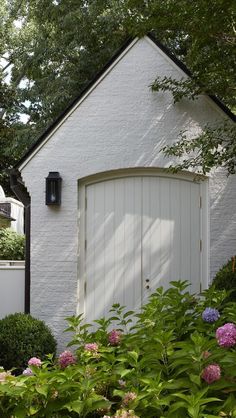 a white building with a door in the middle of some bushes and flowers around it