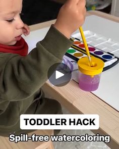 a toddler is playing with paints on a wooden table and holding a pencil in his hand