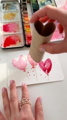 someone is painting hearts on paper with watercolors in the background and another person's hand holding a paintbrush