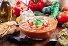 a bowl of tomato soup on a wooden table next to bread, tomatoes and other vegetables