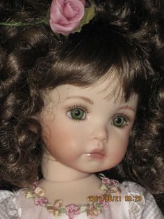 a close up of a doll with curly hair and green eyes wearing a pink flower in her hair