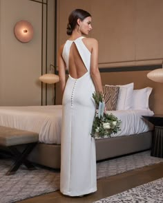 a woman standing in front of a bed wearing a white dress and holding a bouquet