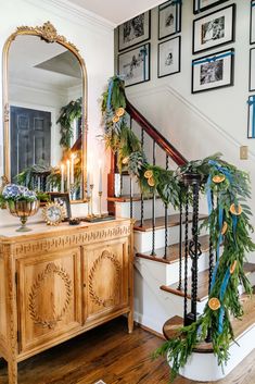 a staircase decorated with greenery next to a wooden dresser and mirror on the wall