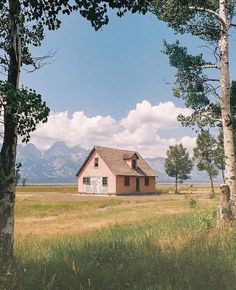 a small house sitting in the middle of a field next to trees and grass with mountains in the background