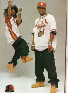 two young men standing next to each other in front of a white background, one holding his hand up