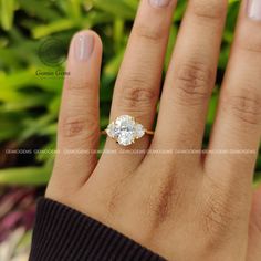 a woman's hand with a diamond ring on top of her finger and green plants in the background