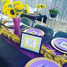 a table set up with purple and yellow plates, napkins and flowers in a vase