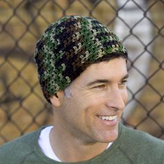 a man wearing a green sweater and a knitted hat smiles at the camera while standing in front of a chain link fence