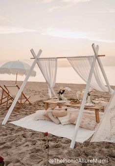 a white canopy bed sitting on top of a sandy beach next to an umbrella covered table