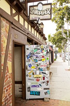 a refrigerator covered in stickers sitting on the side of a building next to a sidewalk