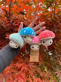 three little crocheted mushrooms are hanging from a tree with autumn leaves in the background