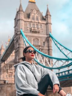 a woman wearing sunglasses sitting on the edge of a bridge in front of a tall building