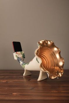 a cell phone in a shell shaped vase on top of a wooden table next to a gold plate