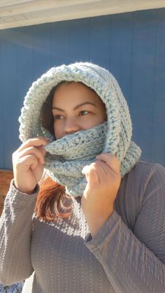 a woman wearing a knitted hood and scarf with her hands under her face while standing in front of a blue wall