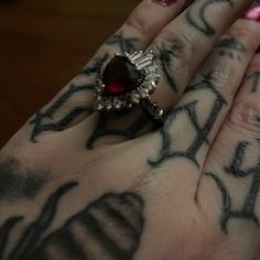 a woman's hand with tattoos and a ring on her left hand, holding a red heart shaped diamond