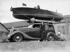 an old black and white photo of a man standing next to a car with a boat on top