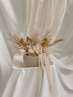 an arrangement of dried flowers in a vase on a white cloth draped chair with drapes