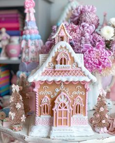 a pink and white gingerbread house on a table next to some flowers in vases