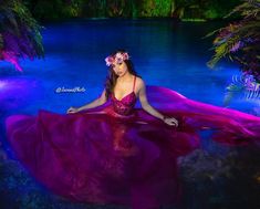 a woman in a long purple dress sitting on the ground next to a body of water