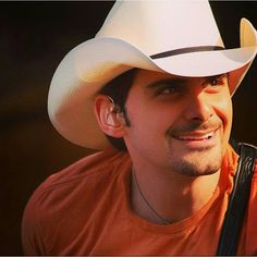 a man in an orange shirt and white cowboy hat smiles at the camera while holding a guitar