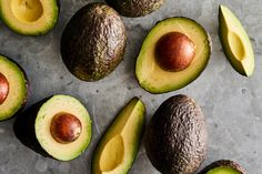 an avocado cut in half and whole on a gray surface with other fruits around it