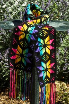 a multicolored knitted shawl with fringes on it in front of purple flowers