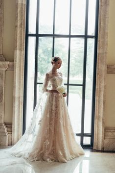 a woman standing in front of a large window wearing a wedding dress with long sleeves