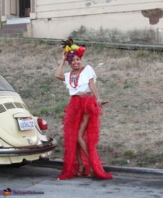 a woman standing next to a car wearing a red skirt and headpiece with flowers in her hair