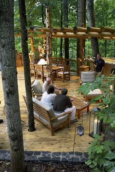 several people sitting on wooden benches in the middle of a wooded area with chairs and tables