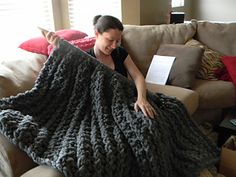 a woman sitting on a couch holding up a large gray crocheted blanket over her