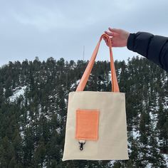 a hand holding an orange and white tote bag in front of snow covered trees