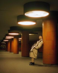 a man leaning against a pillar in an empty building with lights on the ceiling above him