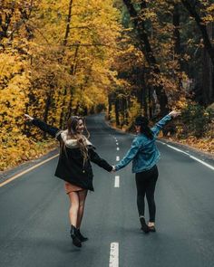 two women holding hands while walking down the road in front of trees with yellow leaves