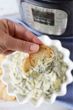 a hand holding a piece of bread over a bowl of spinach artichoke dip