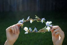 two hands holding up a flower crown in the middle of a grassy area with trees and grass behind them