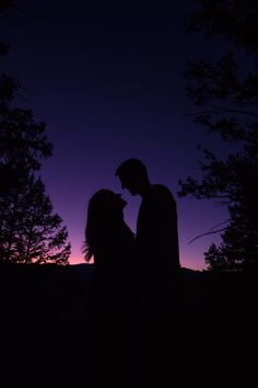 the silhouette of two people standing in front of trees at night, one holding the other's head