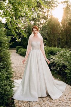 a woman in a wedding dress standing on a gravel path with trees and bushes behind her