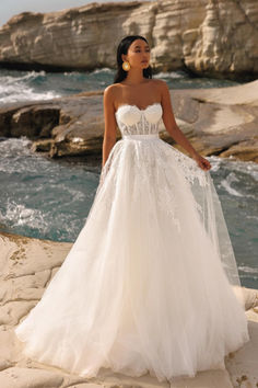 a woman in a white wedding dress standing on the rocks by the ocean with her arms around her waist